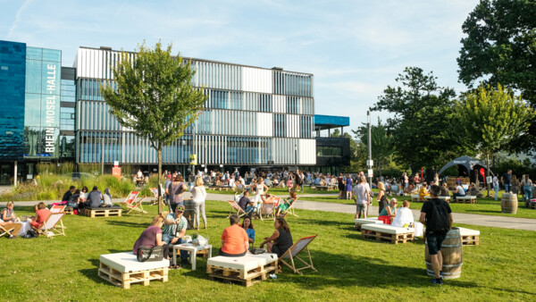 Menschengruppen sitzen verteilt bei der Blaue Stunde vor der Rhein-Mosel-Halle ©Camaze Menschengruppen sitzen verteilt bei der Blaue Stunde vor der Rhein-Mosel-Halle ©Camaze